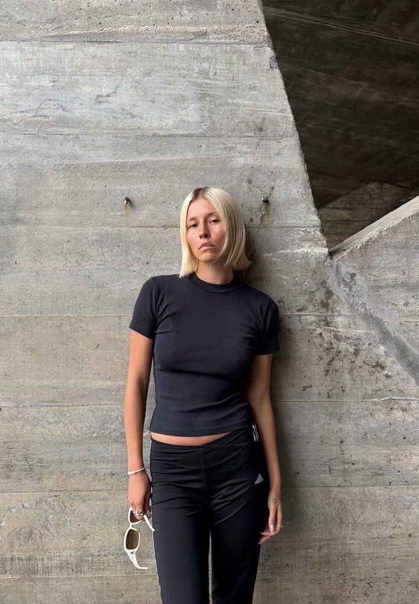 Mujer rubia con camiseta negra de manga corta y pantalones negros Adidas se apoya contra una pared de hormigón rugosa sosteniendo unas gafas de sol blancas en una mano.