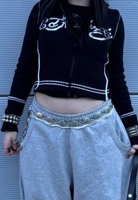 Pull noir court avec des rayures blanches et un design graphique, associé à un pantalon de survêtement gris avec une ceinture argentée décorative et des poches.