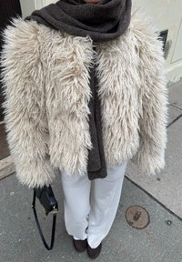 Fluffy kremfarget jakke i imitasjonspels med teksturert overflate, kombinert med et mørkebrunt skjerf. Hvite bukser med vide ben og svart håndveske er synlige.
