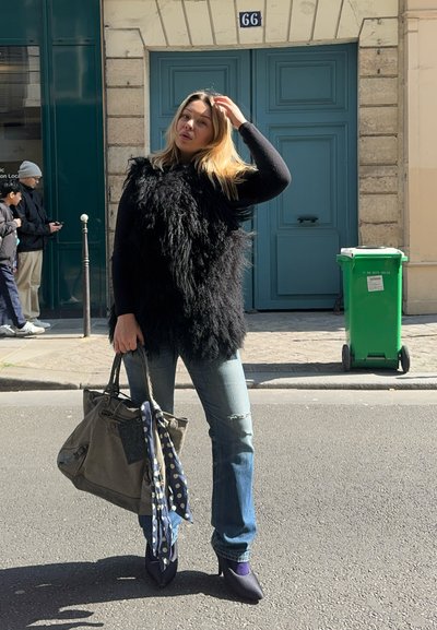 Chaleco de piel sintética negro, blusa negra de mangas largas, jeans desgastados de color azul, tacones morados, llevando un bolso gris con una bufanda de lunares. Fondo de un contenedor verde.