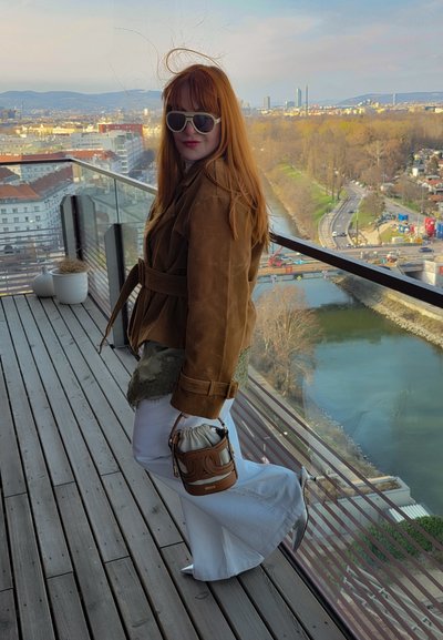 Mujer con cabello rojo que lleva gafas de sol, chaqueta marrón, pantalones blancos y tacones plateados situada en un balcón con vistas al río y al paisaje urbano.