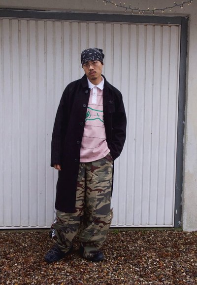 Hombre con bandana negra, gafas, abrigo largo negro, camisa rosa y pantalones de camuflaje, de pie frente a una puerta de garaje blanca sobre suelo de grava.