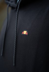 Sudadera negra con capucha que presenta un pequeño logo redondeado en naranja, amarillo y rojo en el lado izquierdo. Incluye capucha y cordones. Textura suave.