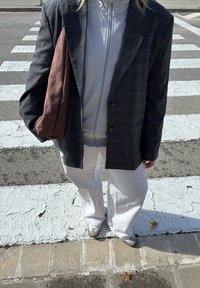 Blazer a quadri grigi sopra una giacca bianca con zip, pantaloni bianchi a gamba larga e sneakers grigi, mentre tiene una borsa in suede marrone. Strisce pedonali visibili sotto.