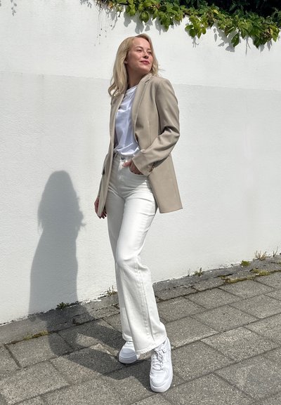 Blazer beige sobre una camiseta blanca, combinado con jeans acampanados blancos y zapatillas blancas. De pie contra una pared blanca con vegetación arriba.