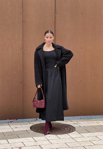 Abrigo negro y texturizado sobre una blusa negra ajustada y una falda larga. Lleva un bolso burdeos. Usa un collar de perlas y botas de tacón burdeos.