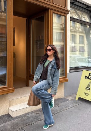 Mujer con chaqueta vaquera y vaqueros, camiseta verde, gafas de sol y zapatillas verdes, de pie sobre una pierna fuera de una tienda con puerta de madera.