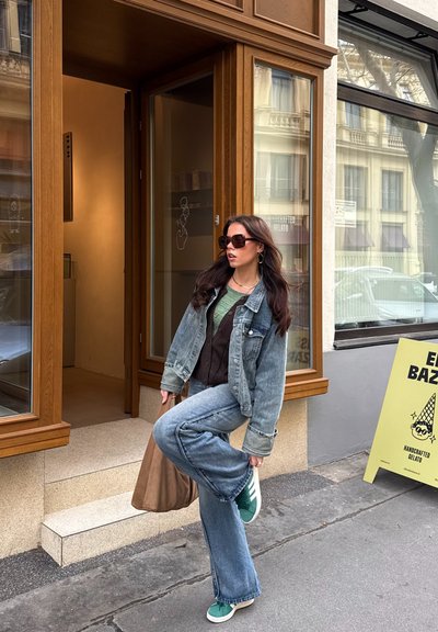 Mujer con chaqueta vaquera y vaqueros, camiseta verde, gafas de sol y zapatillas verdes, de pie sobre una pierna fuera de una tienda con puerta de madera.