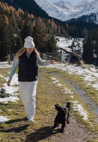 Colete puffer preto sobre um suéter azul claro, calças de fleece de cor creme e um gorro branco. Um cachorro preto corre por um caminho nevado e gramado.
