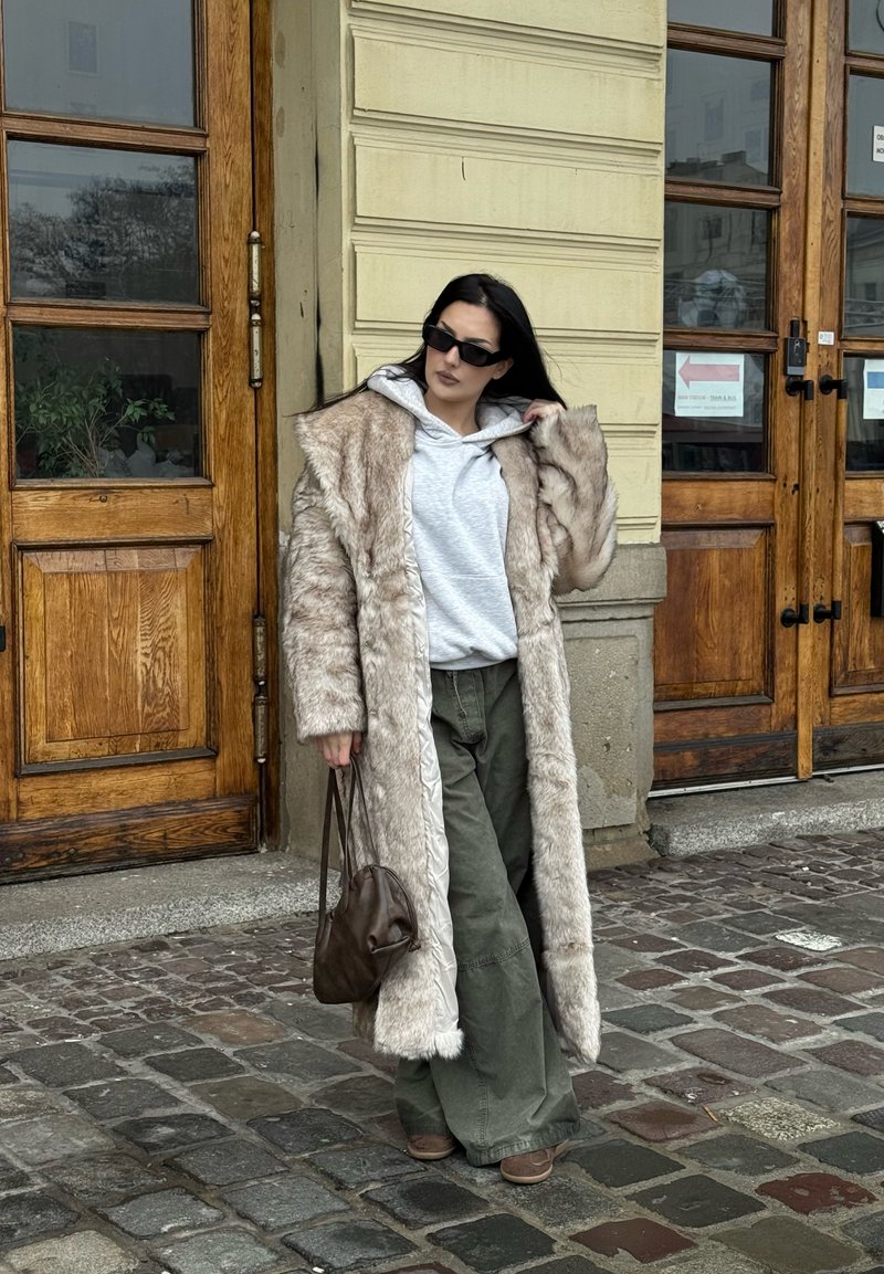 Long manteau en fausse fourrure beige avec de larges revers, sweat à capuche gris, pantalon vert olive, et sac marron. Lunettes de soleil noires complètent le look.