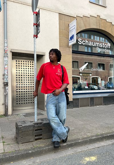 Camisa roja de manga corta con un detalle gráfico, combinada con unos pantalones vaqueros azules holgados. Zapatos de plataforma negros. De pie junto a una señal de tráfico vertical.