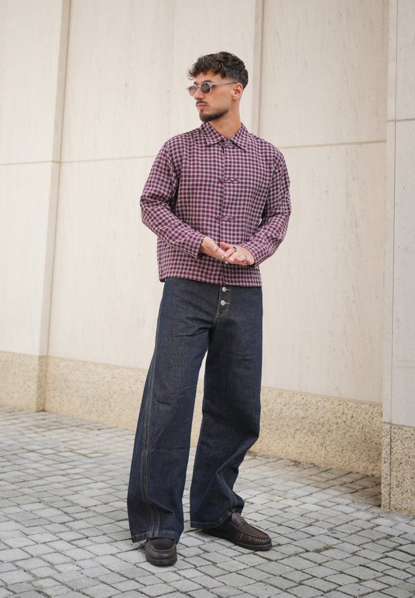 Hombre con camisa de cuadros abotonada, vaqueros anchos y oscuros, gafas de sol y zapatos oscuros, de pie en una calle adoquinada cerca de una pared de piedra clara.