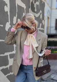 Ljust grå fyrkantig blazer över en rosa tröja, kombinerad med en krämfärgad fluga, blåjeans och en brun mockaväska med silverdetaljer.