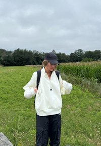 Pullover in pile bianco con colletto zip e dettaglio triangolare nero, abbinato a pantaloni cargo neri e un berretto nero. Sfondo di un campo verde.