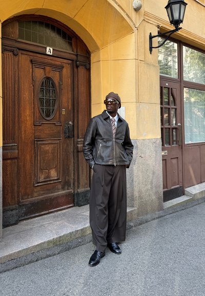 Chaqueta de cuero marrón con bolsillos con cremallera, camisa crema, corbata estampada, pantalones marrones holgados y zapatos negros, frente a una pared amarilla y una puerta de madera.