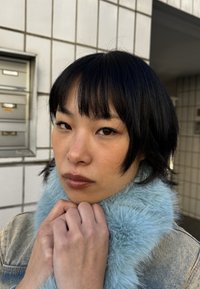 Jeune personne aux cheveux noirs courts portant une veste en jean avec un col en fausse fourrure bleue, tenant le col près contre un mur carrelé en arrière-plan.