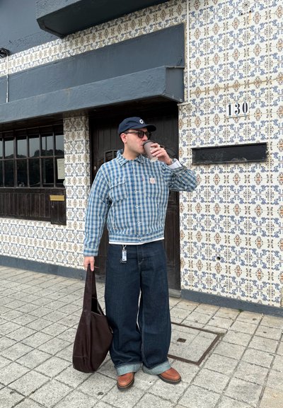 Hombre con chaqueta azul de cuadros, vaqueros anchos y gorra, sosteniendo una bolsa marrón y bebiendo de una taza frente a una pared azulejada con el número 130.