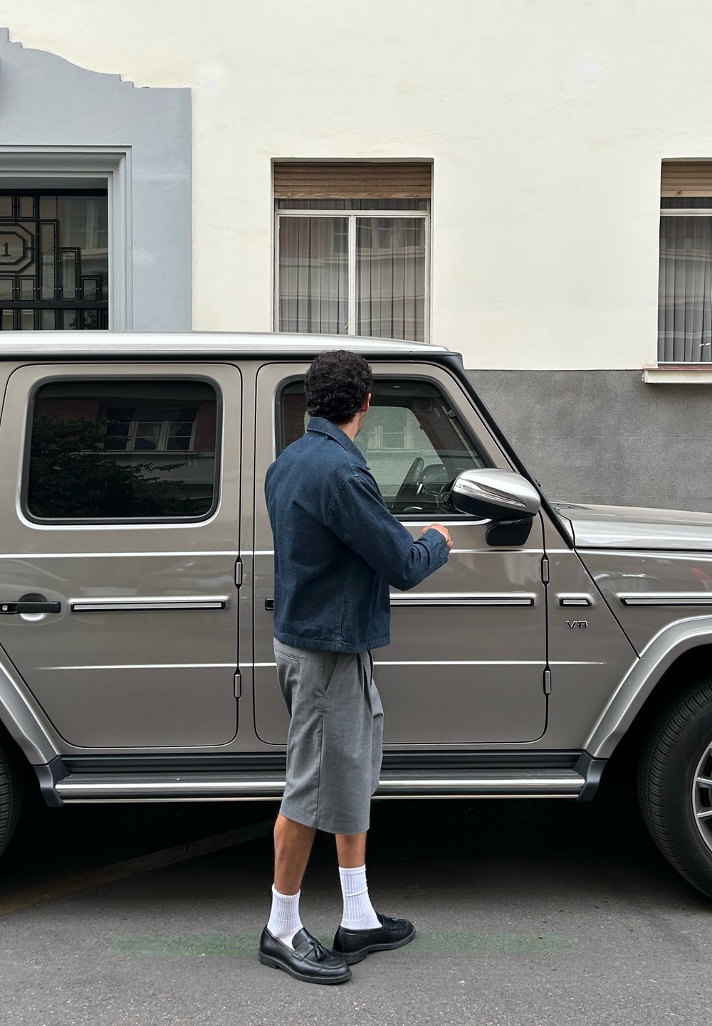 Argent SUV avec une forme rectangulaire, des accents chromés et des fenêtres teintées. Une personne dans une veste bleue et un short gris se tient du côté du conducteur.