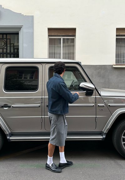 SUV plateado con forma rectangular, acentos de cromo y ventanas oscuras. Persona con chaqueta azul y pantalones cortos grises de pie al lado del conductor.