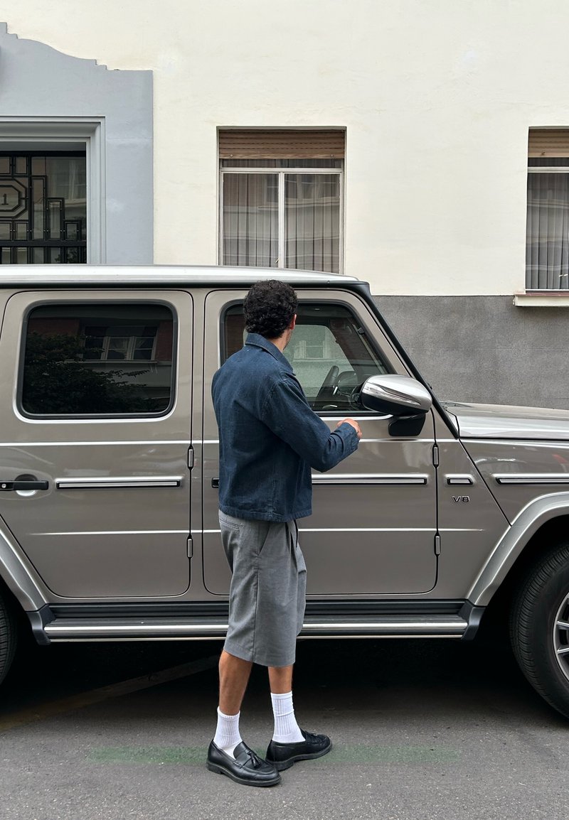 SUV argenté avec une forme rectangulaire, des accents en chrome et des fenêtres teintées. Une personne en veste bleue et en shorts gris se tient du côté conducteur.