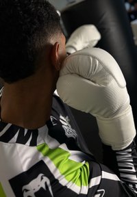 Balti bokso pirštinės su tekstūruota paviršiumi laikomos arti žmogaus veido, kuris dėvi raštuotą juodai žalią treniruočių marškinėlį. Tamsi fono.