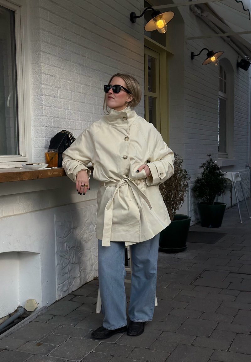 Femme portant un manteau beige ceinturé, des lunettes de soleil et un jean bleu clair, appuyée sur un comptoir extérieur avec une boisson et un sac sous des lampes murales.