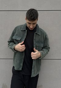 Veste en velours côtelé vert avec un col, deux poches avant et une fermeture à boutons. Portée par-dessus une chemise noire, présentée sur un fond neutre.
