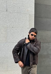 Καφέ δερμάτινο μπουφάν με κοντή σχεδίαση, υφασμάτινη επιφάνεια και μεγάλο γιακά. Συνδυασμένο με μαύρη μπλούζα και γκρίζο παντελόνι.