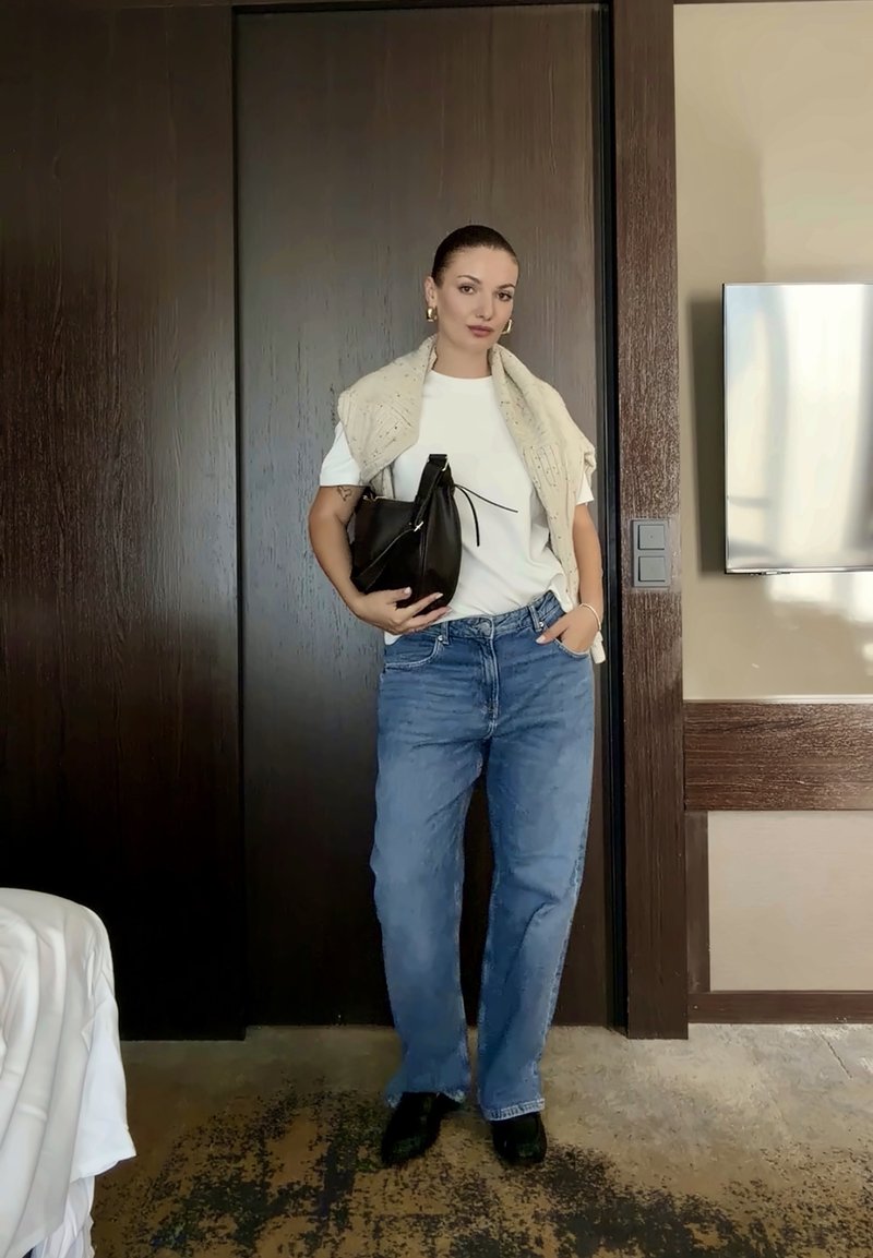 Femme debout à l'intérieur devant des portes en bois foncé, portant une chemise blanche, un pull léger sur les épaules, un jean bleu, des chaussures noires, tenant un sac à main noir.