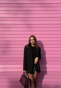 Une femme en blazer noir et jupe tenant un sac à main violet foncé se tient devant un mur rose à panneaux horizontaux.