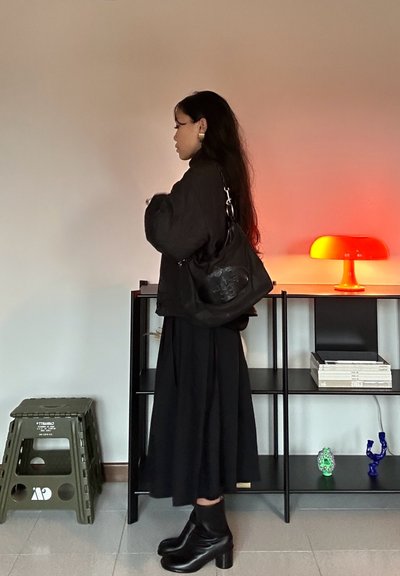 Chaqueta negra acolchada, falda negra fluida y botas negras de tacón grueso. Bolso negro de hombro con detalle en relieve. Lámpara naranja en el fondo.