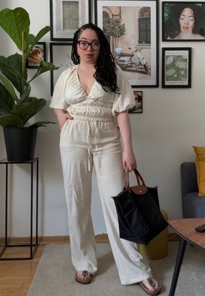 Mono ligero de color crema con escote en V, mangas abullonadas y cinturilla elástica. Bolsa de mano negra con detalles en cuero marrón, muebles de madera.