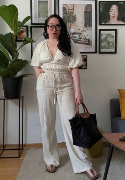 Mono ligero de color crema con escote en V, mangas abullonadas y cinturilla elástica. Bolsa de mano negra con detalles en cuero marrón, muebles de madera.