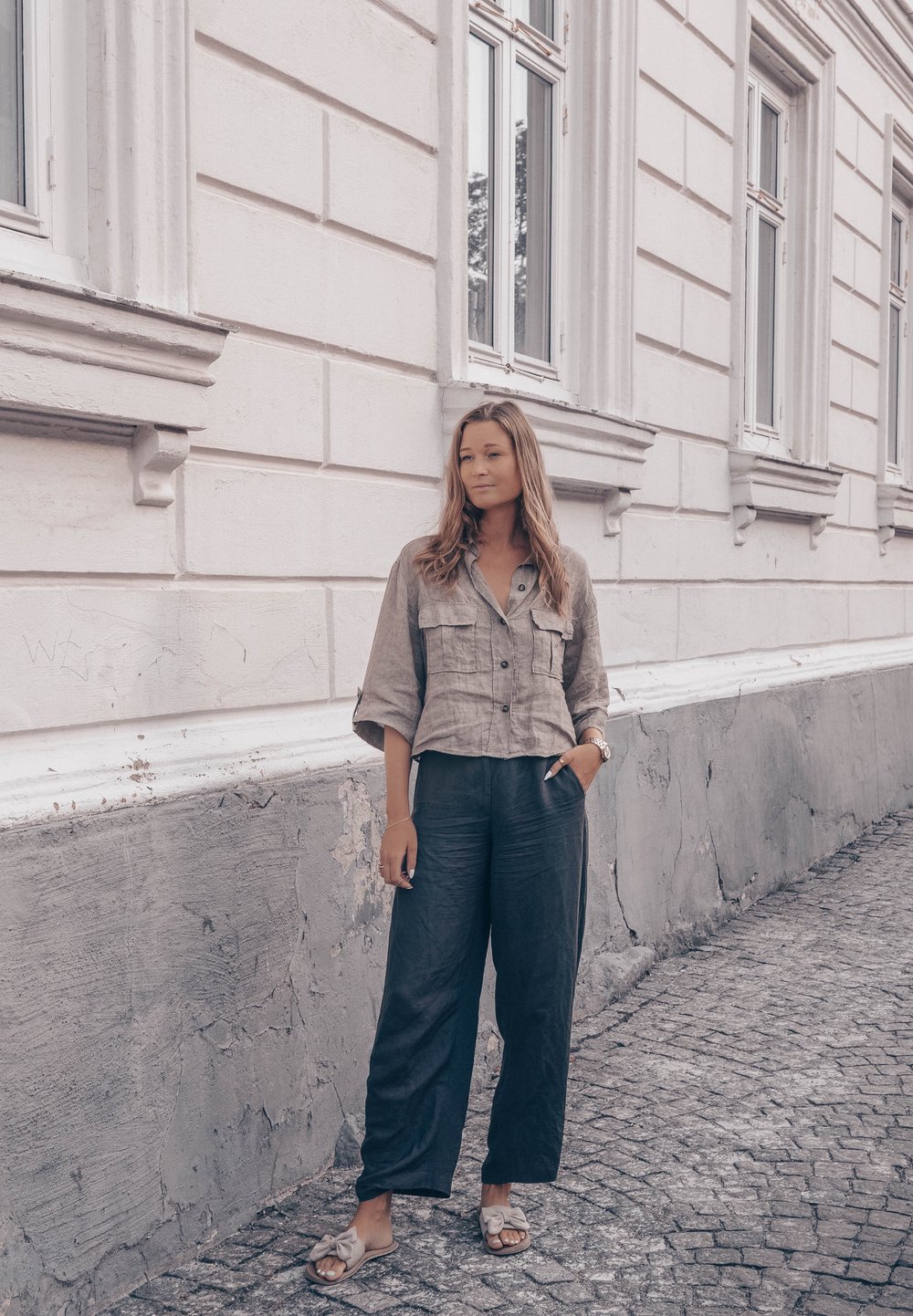 Jasnobrązowa lniana krótka koszula z dwoma kieszeniami na piersi, zestawiona z luźnymi ciemnoszarymi spodniami i beżowymi klapkami. Teksturowane tło.