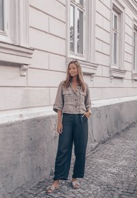 Jasnobrązowa lniana krótka koszula z dwoma kieszeniami na piersi, zestawiona z luźnymi ciemnoszarymi spodniami i beżowymi klapkami. Teksturowane tło.