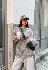 Junge Frau mit rotem Haar, Tarnjacke und Mütze, die eine weiße Kaffeetasse hält, steht auf einem Bürgersteig in der Stadt nahe einer Gebäudeecke.
