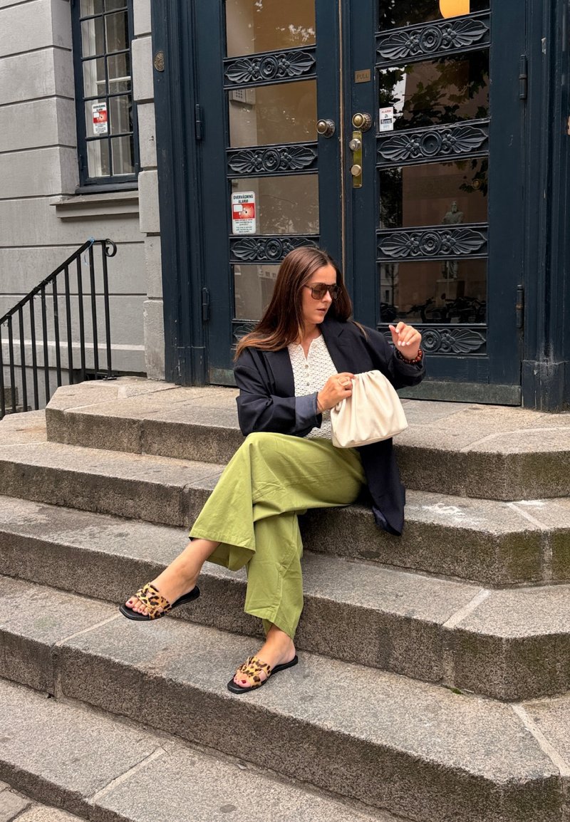 Femme aux longs cheveux bruns portant des lunettes de soleil, un blazer noir, un haut blanc et un pantalon vert, assise sur des marches en pierre en regardant sa montre.