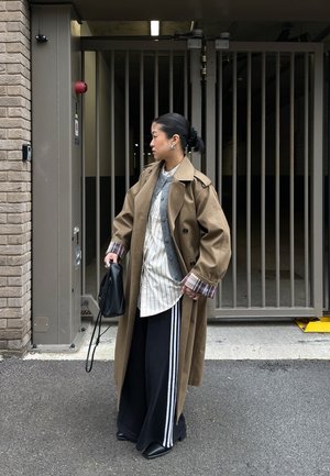 Mujer con gabardina beige sobre camisetas en capas y pantalones anchos negros con rayas blancas, sosteniendo un bolso negro, de pie junto a una puerta metálica cerrada al aire libre.