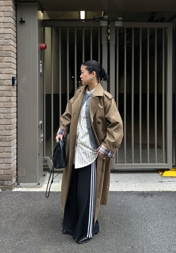 Žena v béžovém trenčkotě přes vrstvené topy a černé široké kalhoty s bílými pruhy, držící černou tašku, stojí venku u zavřené kovové brány.