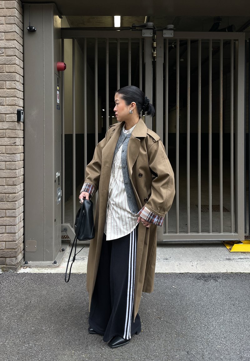 Femme en trench-coat beige portant des hauts superposés et un pantalon large noir avec des rayures blanches, tenant un sac noir, debout près d'un portail métallique fermé en extérieur.