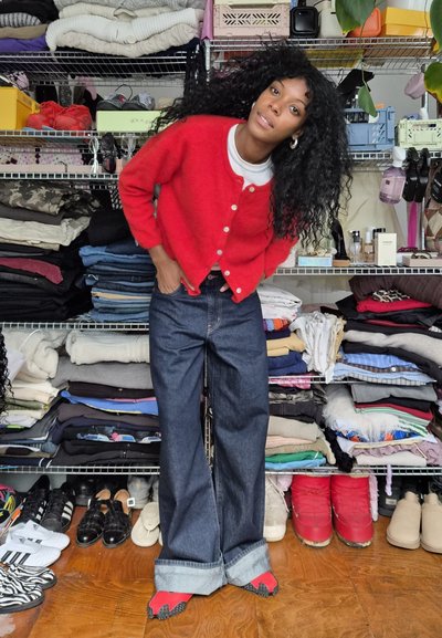 Cárdigan de lana rojo con botones blancos, usado sobre una camiseta blanca. Jeans de mezclilla oscura de pierna ancha y zapatos rojos. Estanterías desordenadas en el fondo.
