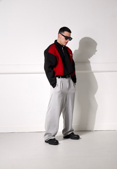 Sudadera oversize roja y negra con cuello alto, pantalones plisados grises y zapatos negros gruesos. Las gafas de sol completan el look.