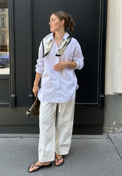 Camisa blanca de botones, de corte holgado, con las mangas arremangadas. Combinada con pantalones anchos blancos y chanclas negras. Bufanda de patrón blanco y negro.