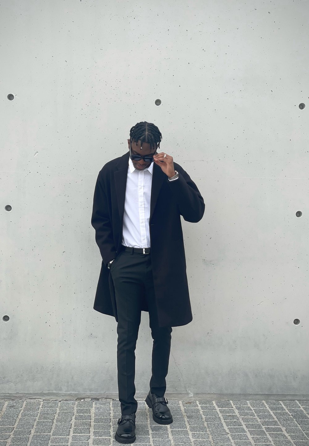 Μαύρο παλτό, λευκό πουκάμισο, μαύρο παντελόνι και μαύρα παπούτσια. Το ντύσιμο διαθέτει κομψό σχέδιο με σύγχρονη γραμμή και μινιμαλιστικά αξεσουάρ.