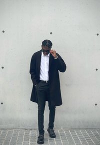 Μαύρο παλτό, λευκό πουκάμισο, μαύρο παντελόνι και μαύρα παπούτσια. Το ντύσιμο διαθέτει κομψό σχέδιο με σύγχρονη γραμμή και μινιμαλιστικά αξεσουάρ.