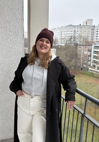 Must pintsak halli kapuutsiga, valged kõrge vöökohaga püksid ja burgundiapunane kootud müts. Naeratab, käed puusal, seisab rõdul.