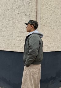 Jeune personne portant une casquette camouflage, un sweat à capuche gris, une veste vert olive et un pantalon beige, debout de profil contre un mur texturé beige et noir.