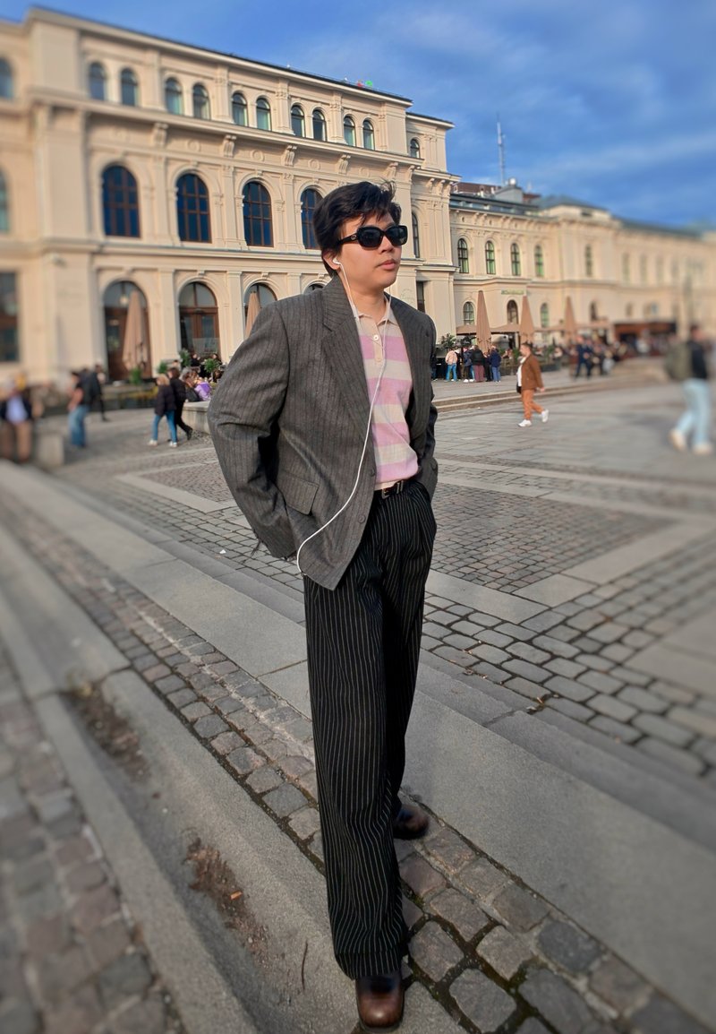 Personne portant des lunettes de soleil, des écouteurs, un blazer gris, une chemise rose rayée et un pantalon noir à fines rayures marchant dans une rue pavée près d'un bâtiment historique.
