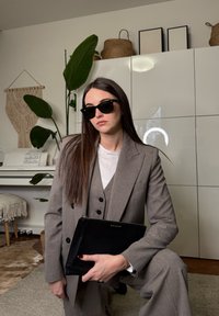 Femme en costume gris et lunettes de soleil tenant une pochette noire, posant à l'intérieur près de plantes, d'un piano et d'une décoration murale tissée.