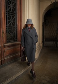 Femme en manteau gris et chapeau assorti marchant devant une porte en bois, portant un sac à main sombre, avec un mur en pierre voûté et une grille en métal à proximité.